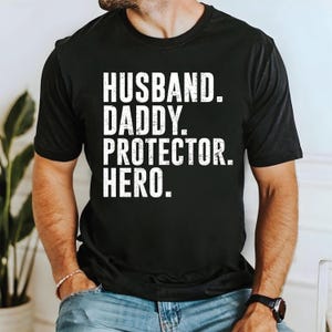Puede incluir: Camiseta negra con texto blanco que dice "Husband. Daddy. Protector. Hero."