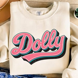 May include: A cream-colored crewneck sweatshirt featuring the word Dolly in a large, stylized pink font with teal and black drop-shadow accents. The garment has long sleeves, a ribbed collar, and a tag at the neckline. The design is centered.