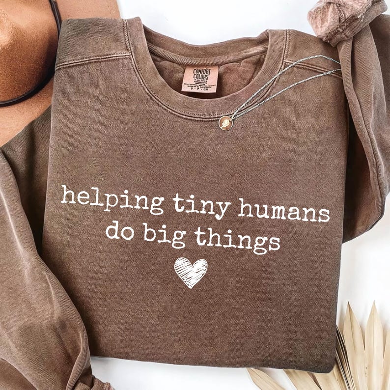 May include: A brown crewneck sweatshirt with a vintage-wash finish. The front features the white text "helping tiny humans do big things" above a small, hand-drawn style white heart. The interior neck label reads "Comfort Colors".