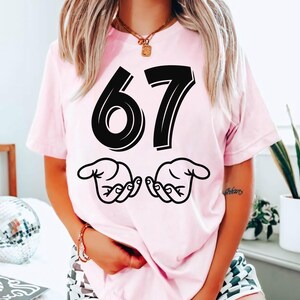 May include: A pale pink t-shirt featuring the number 67 in bold black lettering, with two hand gestures beneath. The t-shirt is a relaxed fit, ideal for casual occasions.