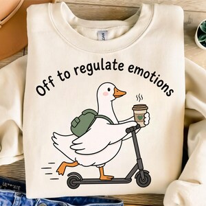 May include: A cream-colored crewneck sweatshirt featuring a graphic of a white duck with an orange beak and feet, wearing a green backpack, riding a black scooter, and holding a coffee cup. The text reads, Off to regulate emotions.
