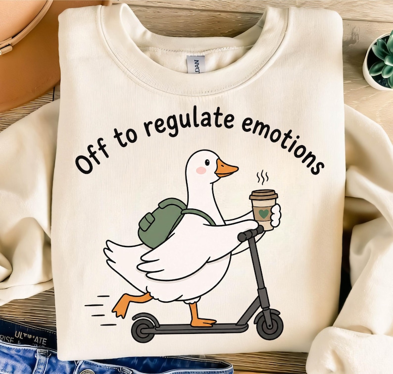 May include: A cream-colored crewneck sweatshirt featuring a graphic of a white duck with an orange beak and feet, wearing a green backpack, riding a black scooter, and holding a coffee cup. The text reads, Off to regulate emotions.