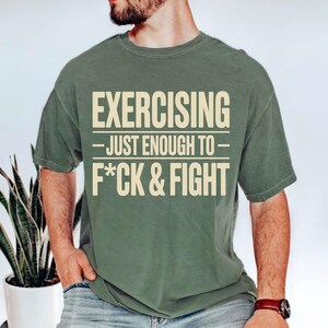 May include: Olive green t-shirt with the text "EXERCISING - JUST ENOUGH TO - F*CK & FIGHT" in a bold, off-white font. The shirt has short sleeves and a crew neck. The shirt is made of a soft, comfortable material.