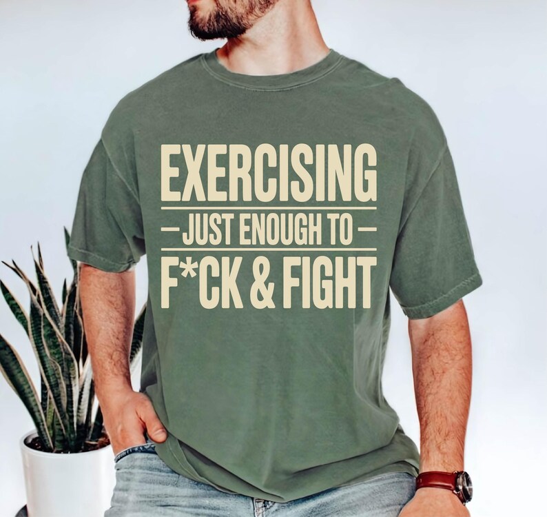 May include: Olive green t-shirt with the text "EXERCISING - JUST ENOUGH TO - F*CK & FIGHT" in a bold, off-white font. The shirt has short sleeves and a crew neck. The shirt is made of a soft, comfortable material.