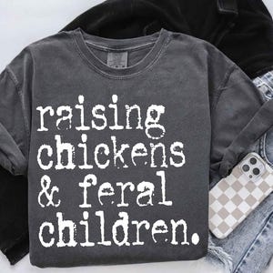 Puede incluir: Una camiseta gris oscura con el texto "raising chickens & feral children." impreso en blanco.