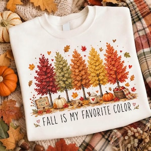 May include: A white crewneck sweatshirt featuring a colorful graphic of five autumn trees in shades of red, orange, and yellow. The design includes pumpkins, a mug, a basket of apples, and the text HELLO FALL and FALL IS MY FAVORITE COLOR.