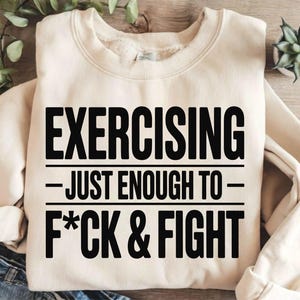 May include: Cream-colored sweatshirt with the text "EXERCISING - JUST ENOUGH TO - F*CK & FIGHT" in bold black letters. The sweatshirt has a crew neck and long sleeves. The background includes greenery and a small plant.