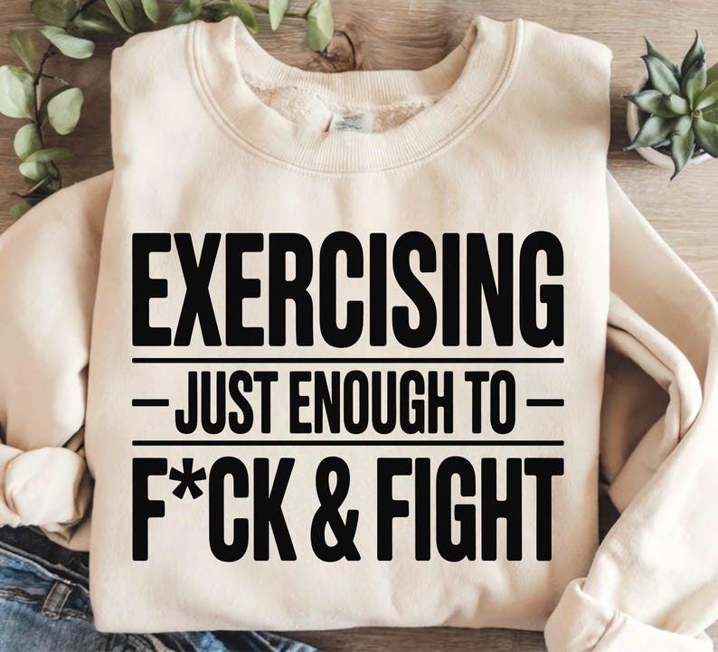 May include: Cream-colored sweatshirt with the text "EXERCISING - JUST ENOUGH TO - F*CK & FIGHT" in bold black letters. The sweatshirt has a crew neck and long sleeves. The background includes greenery and a small plant.