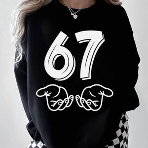 May include: Black sweatshirt featuring the number 67 in a bold, white font. Below the number are two white hand gestures. This casual top is suitable for everyday wear.