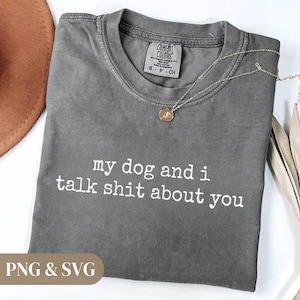 May include: A gray Comfort Colors t-shirt with the text "my dog and i talk shit about you" in white. The shirt has a crew neck and short sleeves. A silver necklace with a small pendant is draped on the shirt.