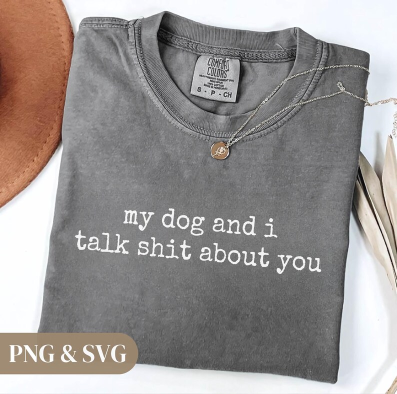 May include: A gray Comfort Colors t-shirt with the text "my dog and i talk shit about you" in white. The shirt has a crew neck and short sleeves. A silver necklace with a small pendant is draped on the shirt.
