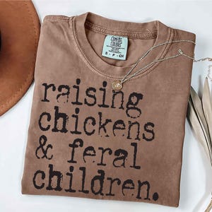 Puede incluir: Una camiseta marr&oacute;n con el texto "raising chickens & feral children." impreso en negro.