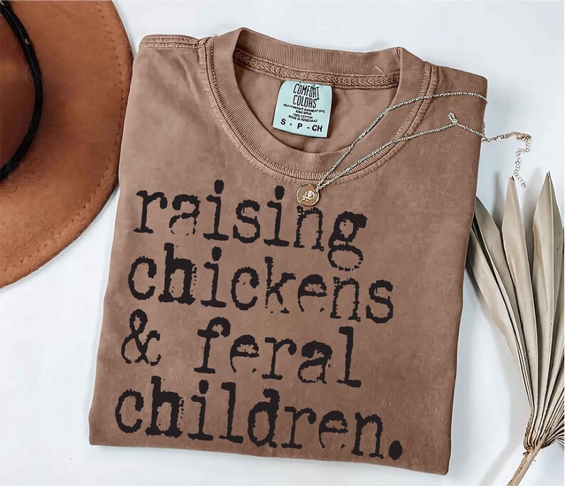 Puede incluir: Una camiseta marr&oacute;n con el texto "raising chickens & feral children." impreso en negro.