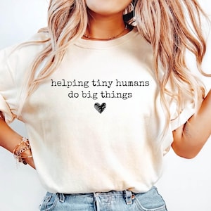 May include: A cream-colored short-sleeve t-shirt featuring the black text "helping tiny humans do big things" printed on the chest, accompanied by a small, hand-drawn style black heart icon centered below the text.