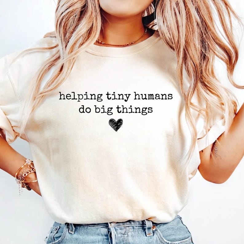 May include: A cream-colored short-sleeve t-shirt featuring the black text "helping tiny humans do big things" printed on the chest, accompanied by a small, hand-drawn style black heart icon centered below the text.