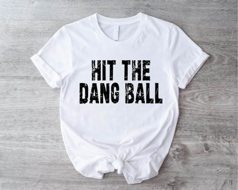 May include: White t-shirt with the phrase "HIT THE DANG BALL" printed in distressed black letters. The shirt is made of a soft material and has short sleeves. The shirt is knotted at the waist.
