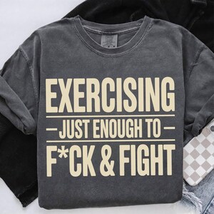 May include: Dark gray sweatshirt with the text "EXERCISING - JUST ENOUGH TO - F*CK & FIGHT" in a cream-colored font. The sweatshirt is folded on a white surface with a phone case visible.