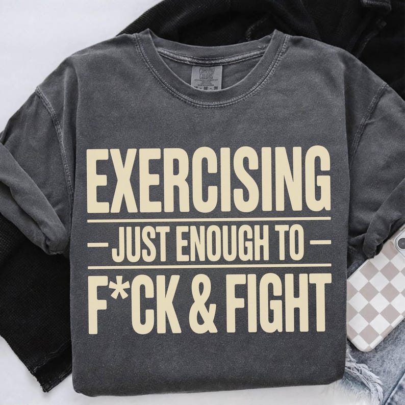 May include: Dark gray sweatshirt with the text "EXERCISING - JUST ENOUGH TO - F*CK & FIGHT" in a cream-colored font. The sweatshirt is folded on a white surface with a phone case visible.