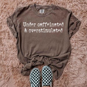 Puede incluir: Una camiseta marrón con el texto "Under caffeinated & overstimulated" impreso en blanco.