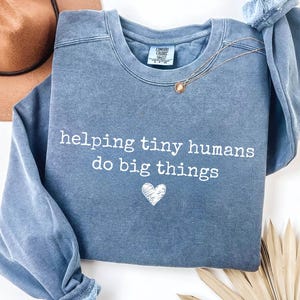 May include: A blue crewneck sweatshirt with a washed-out finish featuring the white text "helping tiny humans do big things" and a small white heart graphic centered below the text. The garment has a ribbed collar and long sleeves.