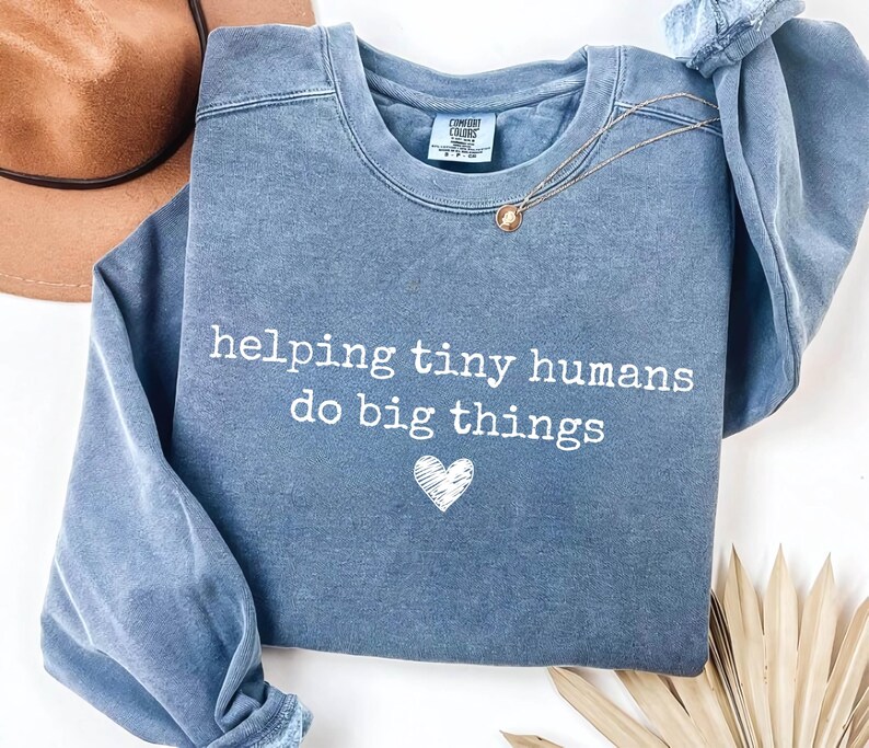 May include: A blue crewneck sweatshirt with a washed-out finish featuring the white text "helping tiny humans do big things" and a small white heart graphic centered below the text. The garment has a ribbed collar and long sleeves.