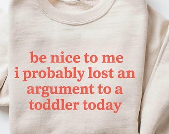 Op de afbeelding: Beige sweatshirt met de rode tekst: "be nice to me i probably lost an argument to a toddler today". De sweatshirt heeft een ronde hals en lange mouwen. De tekst is in een vetgedrukte, schreefloze lettertype.