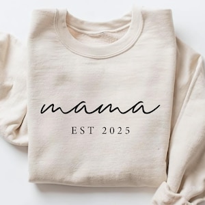 Puede incluir: Una sudadera de cuello redondo color crema con la palabra mama en una fuente cursiva negra centrada en el pecho. Debajo de la escritura, el texto EST 2025 está impreso en una fuente serif negra sencilla. La prenda tiene mangas largas y cuello acanalado.