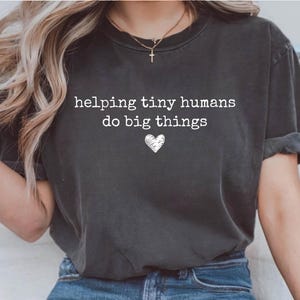 May include: A charcoal gray short-sleeve t-shirt featuring the white text "helping tiny humans do big things" printed on the chest, accompanied by a small white hand-drawn heart icon centered below the text.