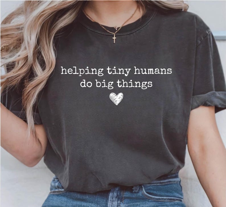 May include: A charcoal gray short-sleeve t-shirt featuring the white text "helping tiny humans do big things" printed on the chest, accompanied by a small white hand-drawn heart icon centered below the text.
