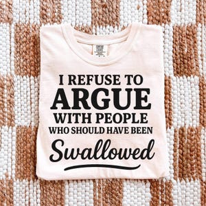 K&ouml;nnte beinhalten: Hellpfirsichfarbenes T-Shirt mit schwarzem Text "I REFUSE TO ARGUE WITH PEOPLE WHO SHOULD HAVE BEEN SWALLOWED". Das T-Shirt hat einen Rundhalsausschnitt und ist aus weichem Material.
