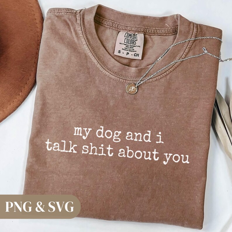 May include: A brown Comfort Colors t-shirt with the text "my dog and i talk shit about you" in white. A silver necklace with a pendant is on the shirt. The shirt is folded on a white surface.