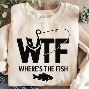May include: A cream-coloured sweatshirt with the text "WTF WHERE'S THE FISH" in black, featuring a fishing hook and a fish graphic. This casual garment is ideal for those who enjoy fishing.