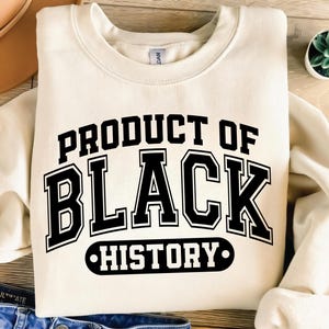 Peut inclure: Sweat-shirt crème avec l'inscription "PRODUCT OF BLACK HISTORY" en lettres noires et grasses de style universitaire. Le sweat-shirt est plié, avec une petite plante succulente dans un pot blanc en arrière-plan.
