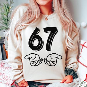 May include: A beige sweatshirt featuring the number "67" in a bold black font, and two hand illustrations. The design is printed on a casual sweatshirt, suitable for everyday wear.