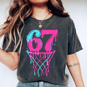 May include: Dark grey t-shirt with a basketball-themed graphic. The design features the number "67" in pink with blue dripping accents, above a basketball net. The net and dripping accents are in blue and pink.