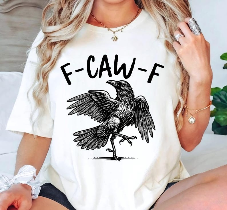 May include: Cream-colored t-shirt featuring a black graphic of a crow with outstretched wings and the text "F-CAW-F" above it. The crow is depicted in a detailed, illustrative style. The shirt is a casual wear item.