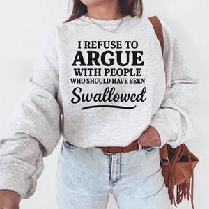 K&ouml;nnte beinhalten: Hellgraues Sweatshirt mit dem schwarzen Text: "I REFUSE TO ARGUE WITH PEOPLE WHO SHOULD HAVE BEEN SWALLOWED". Das Sweatshirt hat lange &Auml;rmel und einen Rundhalsausschnitt. Die Person tr&auml;gt eine blaue Jeans und einen braunen G&uuml;rtel.