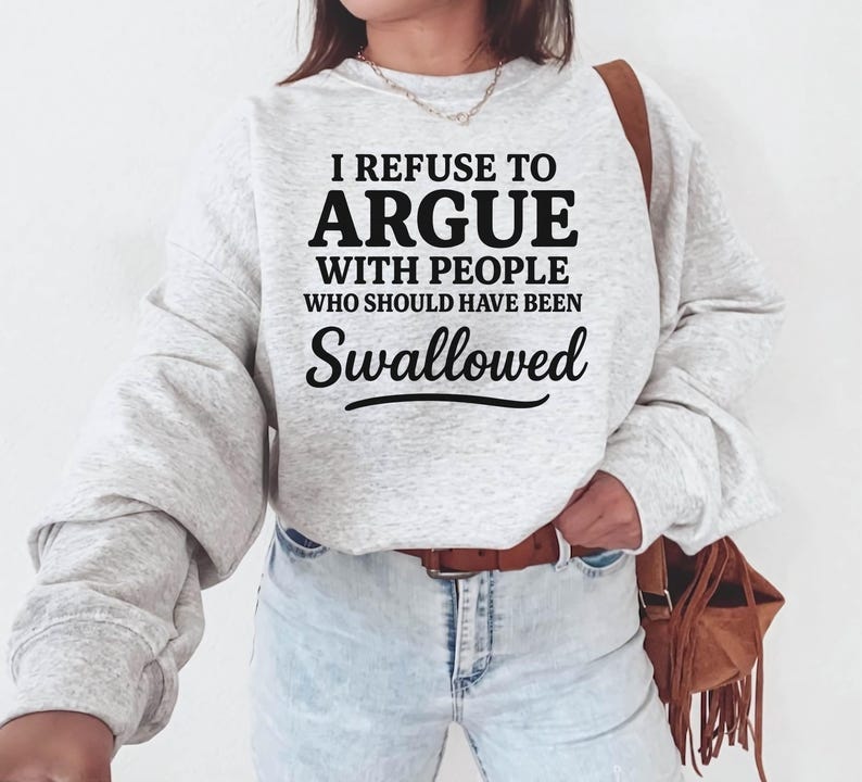 K&ouml;nnte beinhalten: Hellgraues Sweatshirt mit dem schwarzen Text: "I REFUSE TO ARGUE WITH PEOPLE WHO SHOULD HAVE BEEN SWALLOWED". Das Sweatshirt hat lange &Auml;rmel und einen Rundhalsausschnitt. Die Person tr&auml;gt eine blaue Jeans und einen braunen G&uuml;rtel.