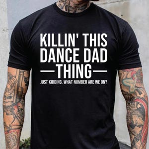 Può includere: T-shirt nero con testo bianco "KILLIN' THIS DANCE DAD THING" e la scritta "JUST KIDDING. WHAT NUMBER ARE WE ON?" sotto. La maglietta è a girocollo.
