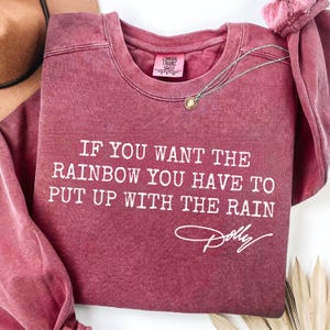 May include: A maroon long-sleeve crewneck sweatshirt with a vintage-wash finish. The front features white text that reads, "IF YOU WANT THE RAINBOW YOU HAVE TO PUT UP WITH THE RAIN" followed by a white script signature that reads, "Dolly".