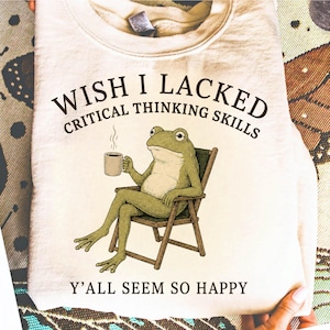 Pode incluir: Uma camisola de gola redonda cor de creme com o gráfico de um sapo verde sentado numa cadeira de madeira enquanto segura uma caneca de café a fumegar. O texto acima diz WISH I LACKED CRITICAL THINKING SKILLS e abaixo Y'ALL SEEM SO HAPPY.