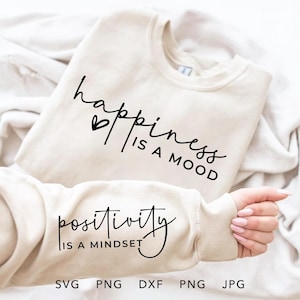 May include: A white sweatshirt with the text "happiness is a mood" and "positivity is a mindset" printed on it. The sweatshirt is folded and lying on a white surface.