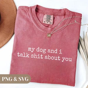 May include: A dusty rose-colored t-shirt with the text "my dog and i talk shit about you" in white. A silver necklace with a small pendant rests on the shirt. The shirt has a Comfort Colors tag.