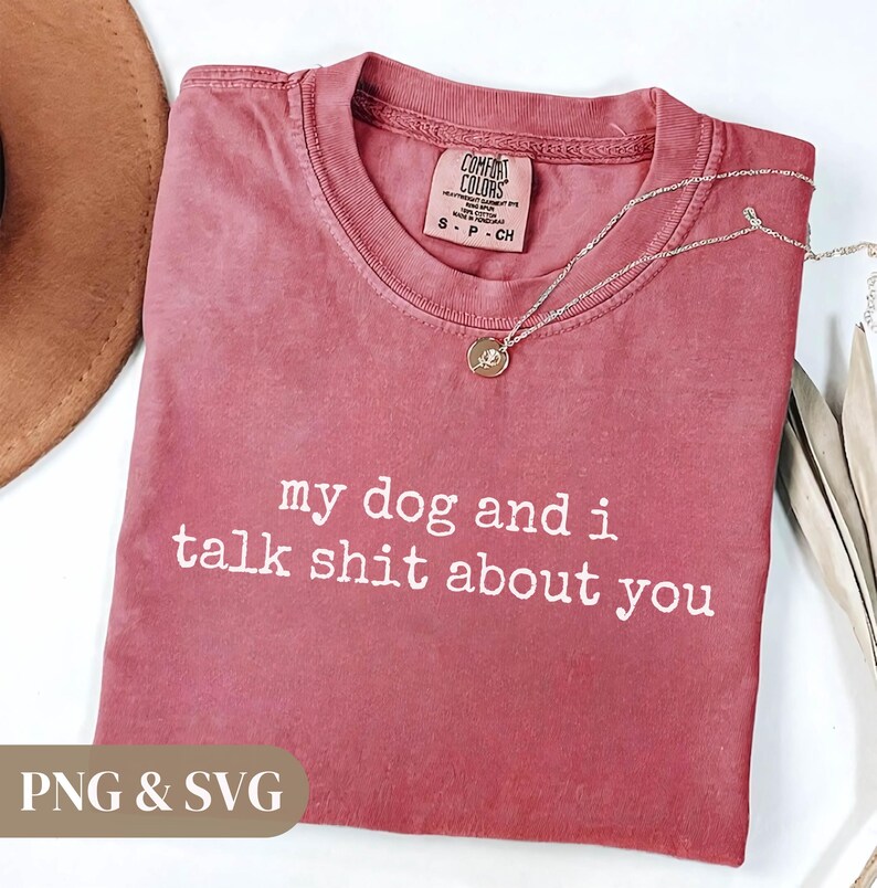 May include: A dusty rose-colored t-shirt with the text "my dog and i talk shit about you" in white. A silver necklace with a small pendant rests on the shirt. The shirt has a Comfort Colors tag.