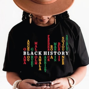 Peut inclure: T-shirt noir avec l'inscription "BLACK HISTORY" en blanc, avec les noms de personnalités historiques en vert, rouge et jaune. La personne sur l'image porte un chapeau marron et tient un téléphone.