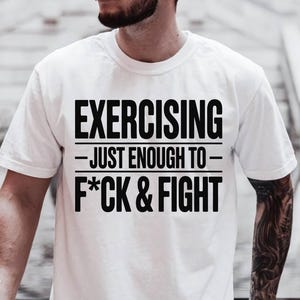 May include: White t-shirt with the text "EXERCISING - JUST ENOUGH TO - F*CK & FIGHT" in bold black letters. The shirt has a crew neck and short sleeves. The image is a close-up of the shirt.
