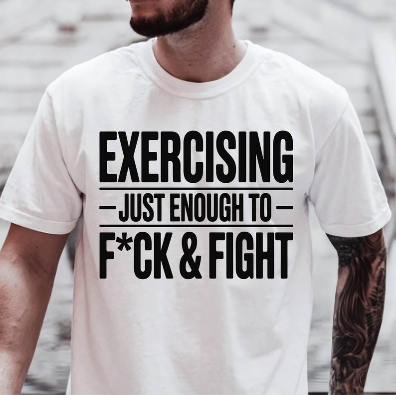 May include: White t-shirt with the text "EXERCISING - JUST ENOUGH TO - F*CK & FIGHT" in bold black letters. The shirt has a crew neck and short sleeves. The image is a close-up of the shirt.