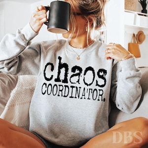 May include: A light gray sweatshirt with the words "chaos COORDINATOR" printed in black. The person in the image is holding a black mug. The sweatshirt is a casual style.
