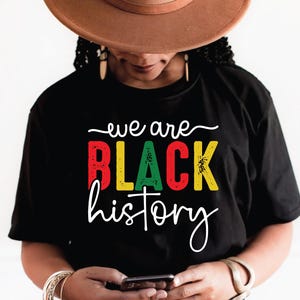 Może przedstawiać: Czarny t-shirt z napisem "we are BLACK history" w kolorze białym, czerwonym, zielonym i żółtym. Koszulka jest noszona przez osobę w brązowym kapeluszu.
