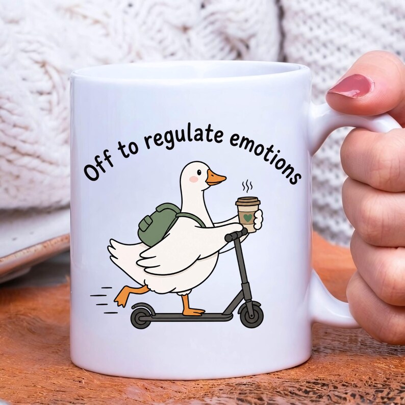 May include: A white ceramic mug featuring a cartoon illustration of a white duck with an orange beak and feet, wearing a green backpack, riding a black scooter, and holding a coffee cup. The text "Off to regulate emotions" is printed above.